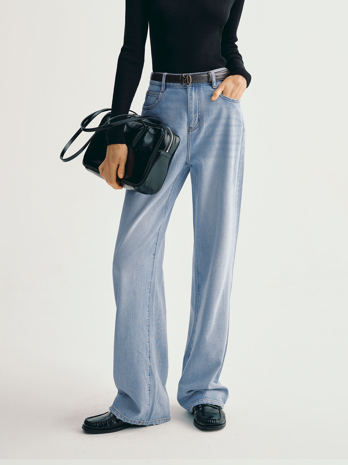 Person wearing light blue jeans and black shoes, holding a black handbag on a white background