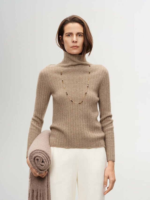 Woman wearing a beige turtleneck sweater holding a folded blanket against a plain background