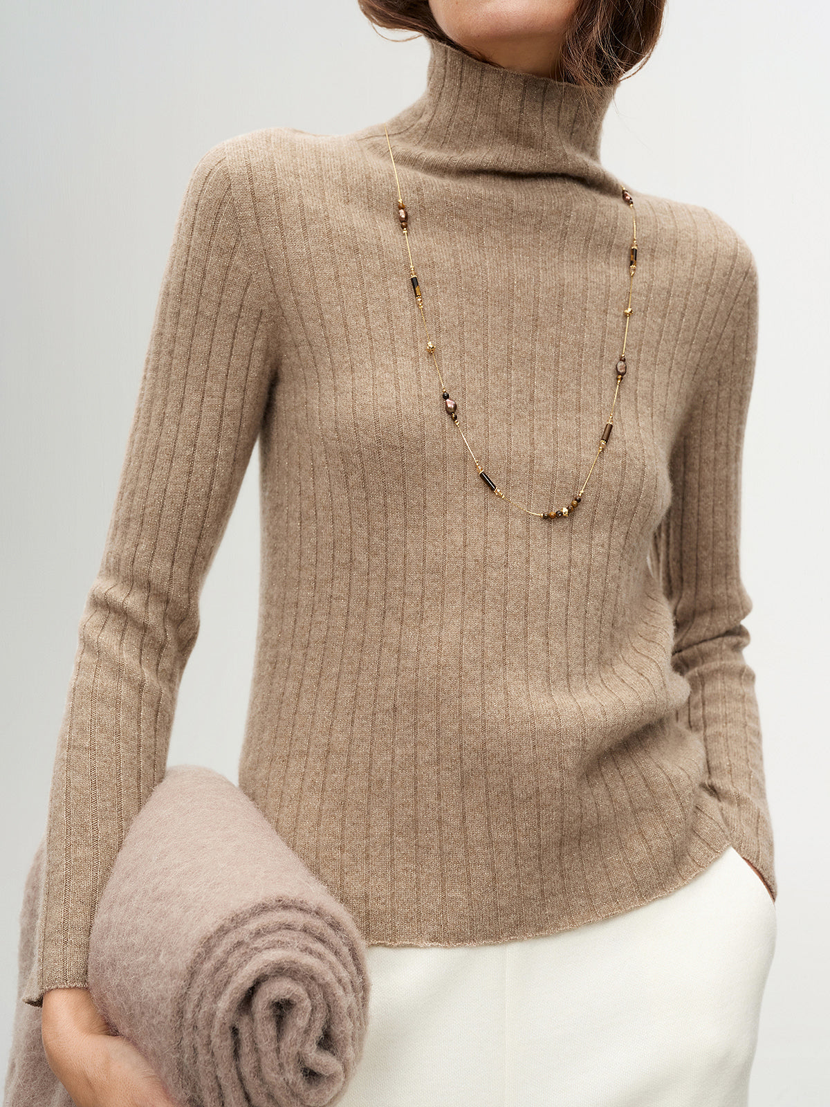 Person wearing a beige ribbed turtleneck sweater holding a folded blanket against a plain background