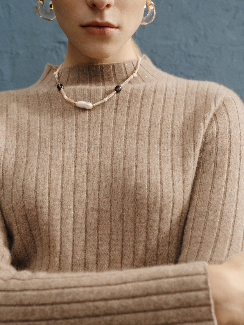 Person wearing a beige ribbed sweater and a beaded necklace against a blue background
