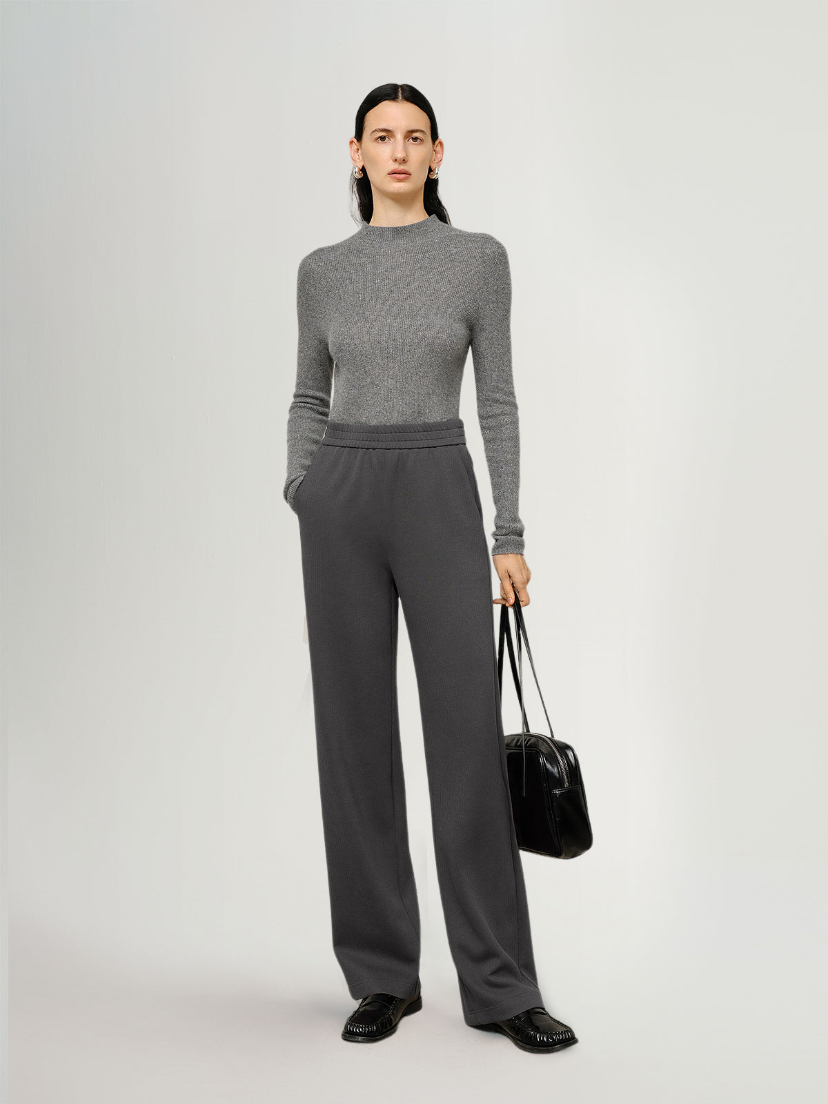 Person wearing a gray sweater and pants holding a black bag on a white background