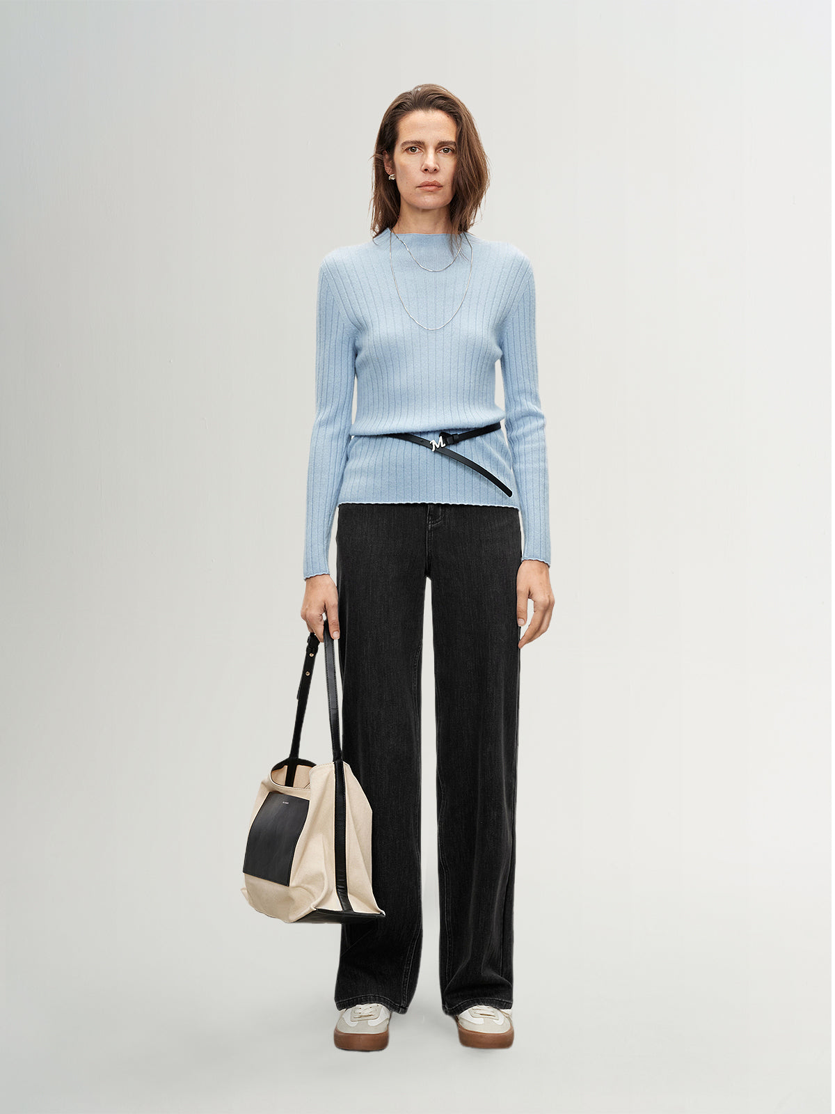 Woman wearing a light blue sweater and black pants, holding a beige and black bag on a white background