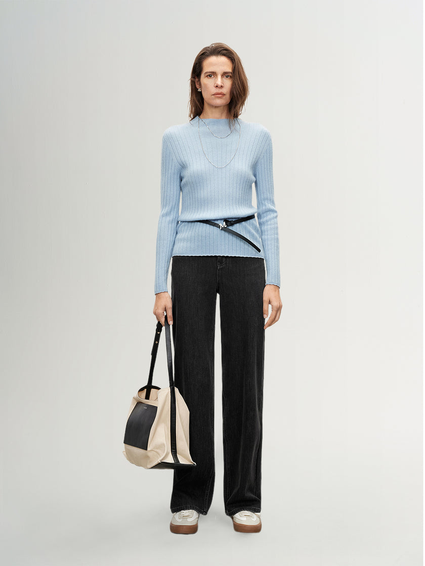 Woman wearing a light blue sweater and black pants, holding a beige and black bag on a white background