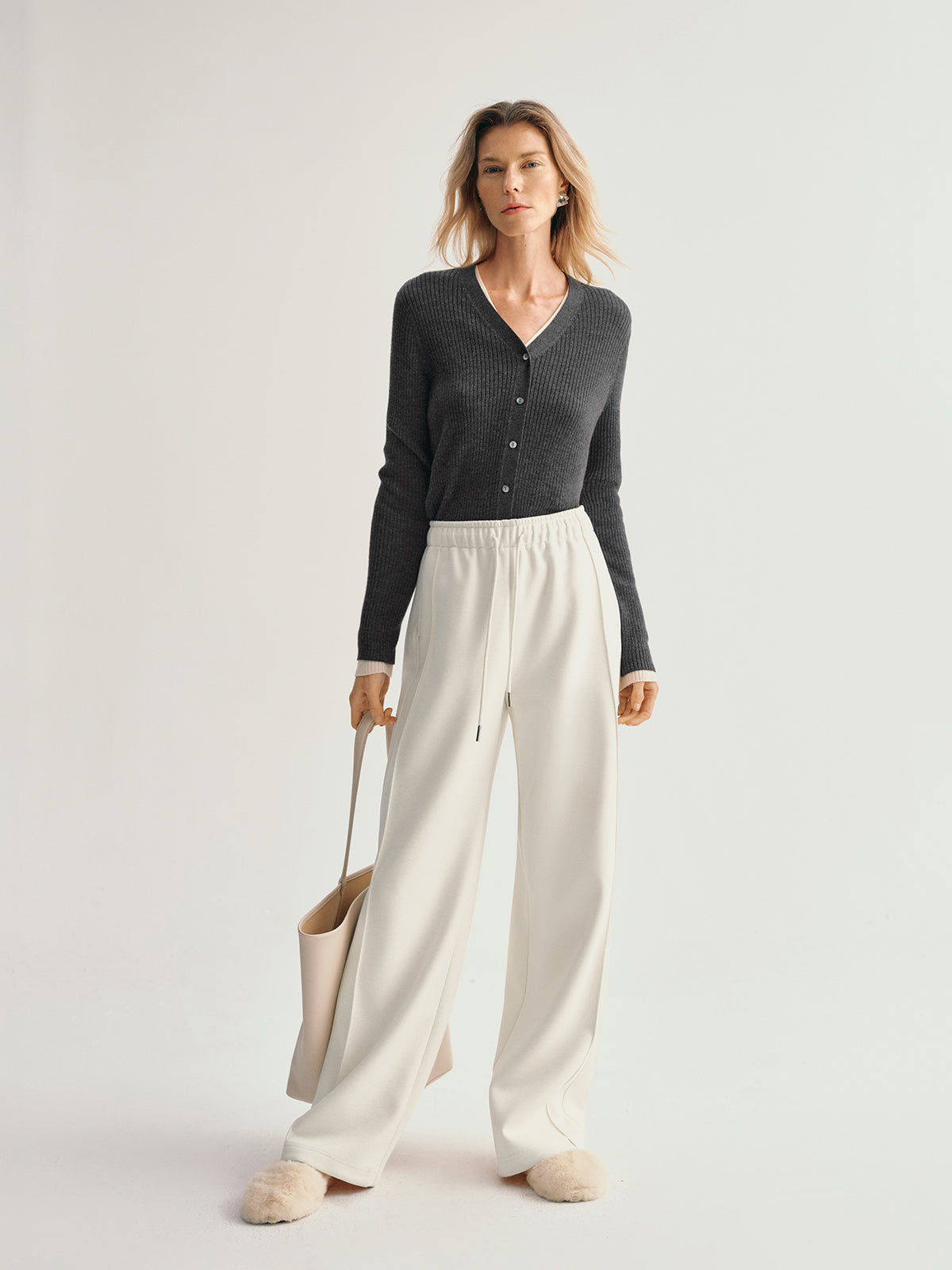 Woman wearing a dark gray cardigan and white pants holding a beige bag on a light background