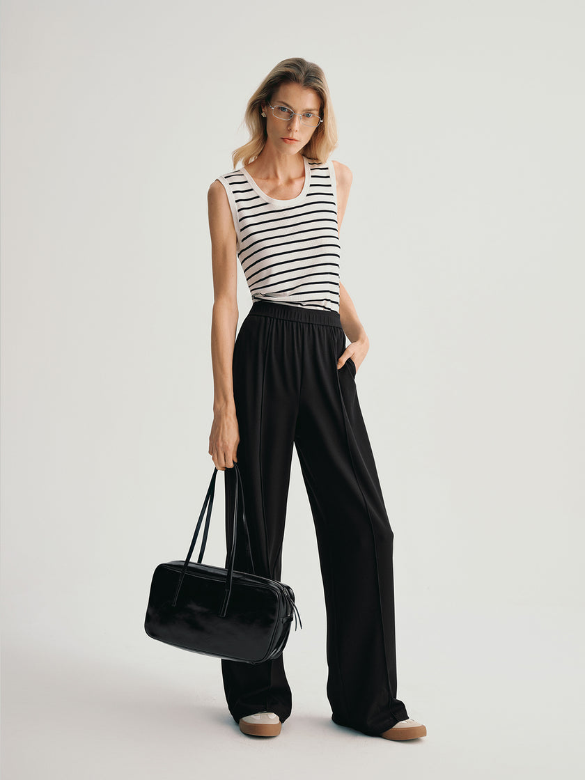 Woman wearing a black and white striped sleeveless top and black wide-leg pants, holding a black handbag on a light gray background.