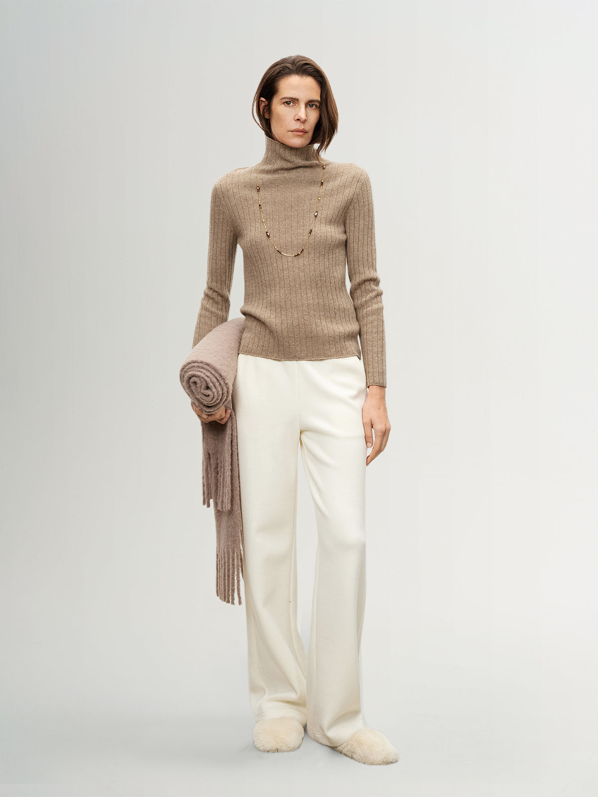 Woman wearing a beige turtleneck sweater and white pants, holding a beige scarf.