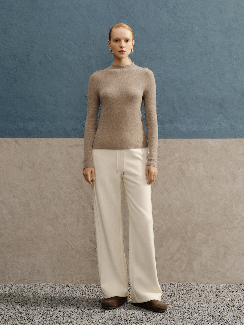 Person wearing a beige sweater and white pants against a blue and gray background