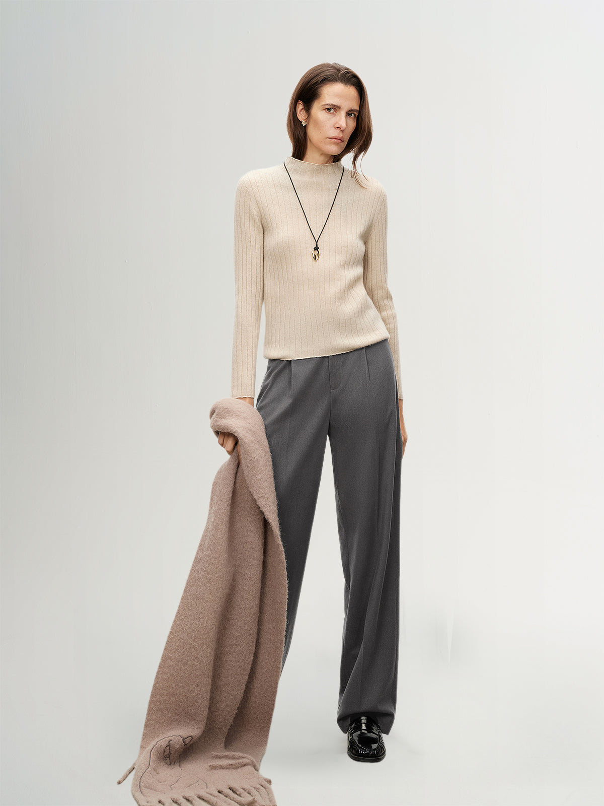 Woman wearing a beige sweater and gray pants holding a brown scarf on a white background