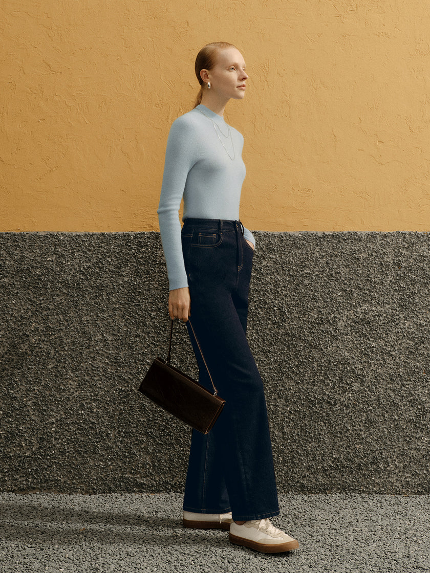 Woman in a light gray turtleneck and dark blue jeans standing against a yellow and gray wall.