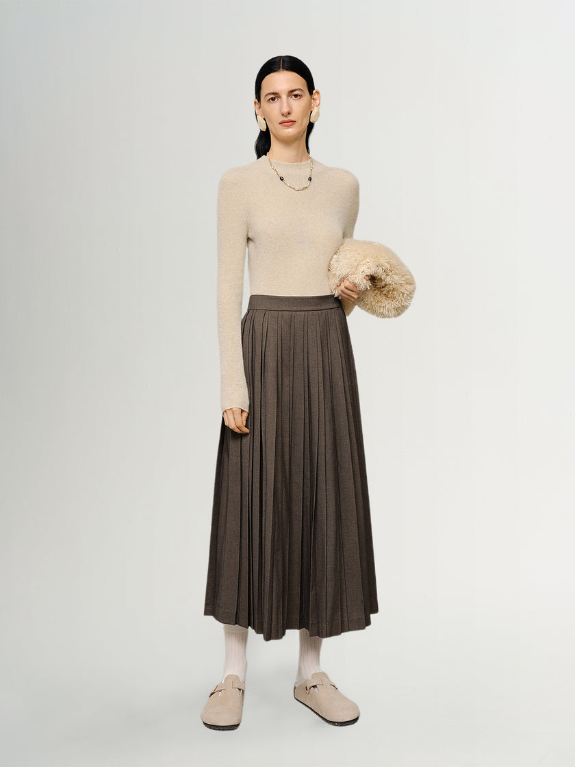 Woman wearing a beige sweater and brown pleated skirt holding a beige bag on a white background