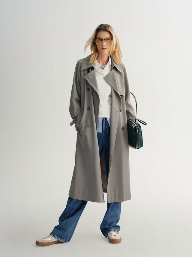A woman modeling a mid-length gray trench coat with a double-breasted design and a notch lapel collar. She is also wearing blue jeans and carrying a black bag.