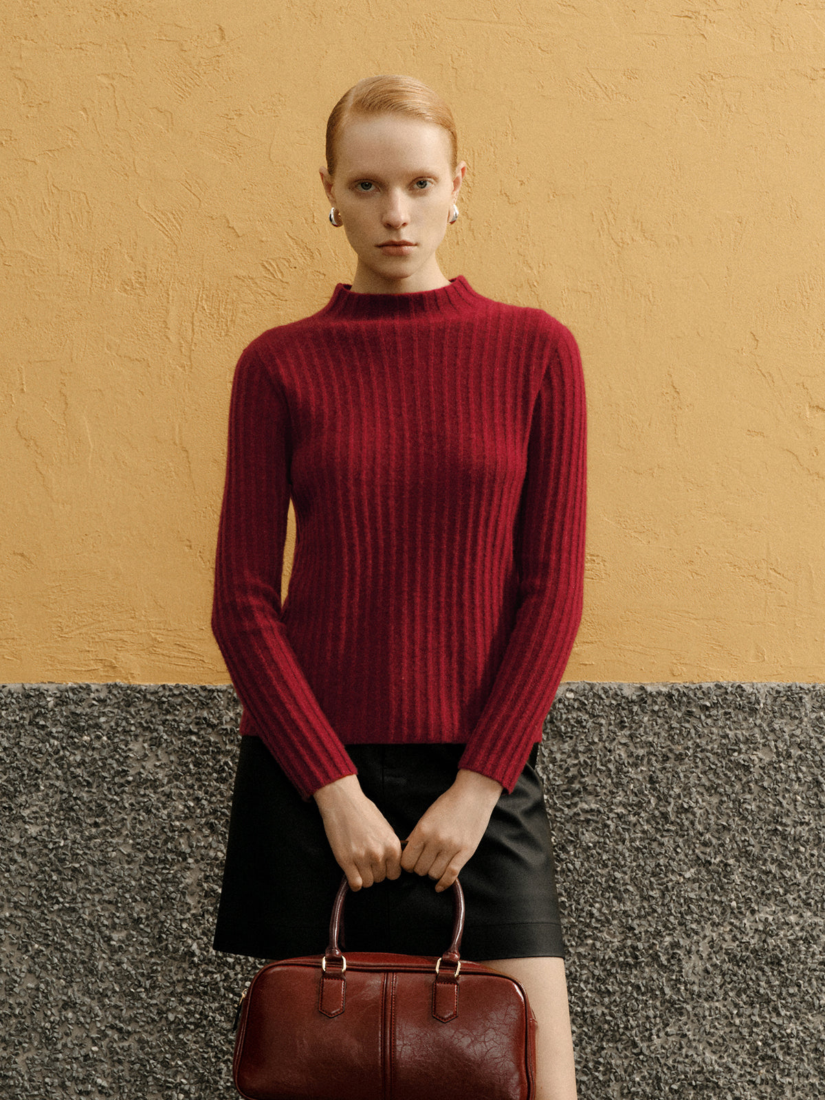Woman wearing a red sweater and black skirt holding a brown handbag against a yellow and gray background
