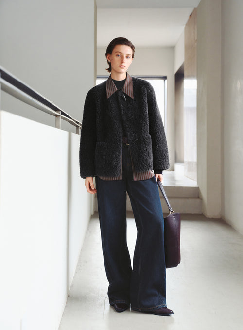 Load image 1 in gallery view. Person wearing a dark textured jacket and blue pants, holding a purple bag in a minimalistic setting.