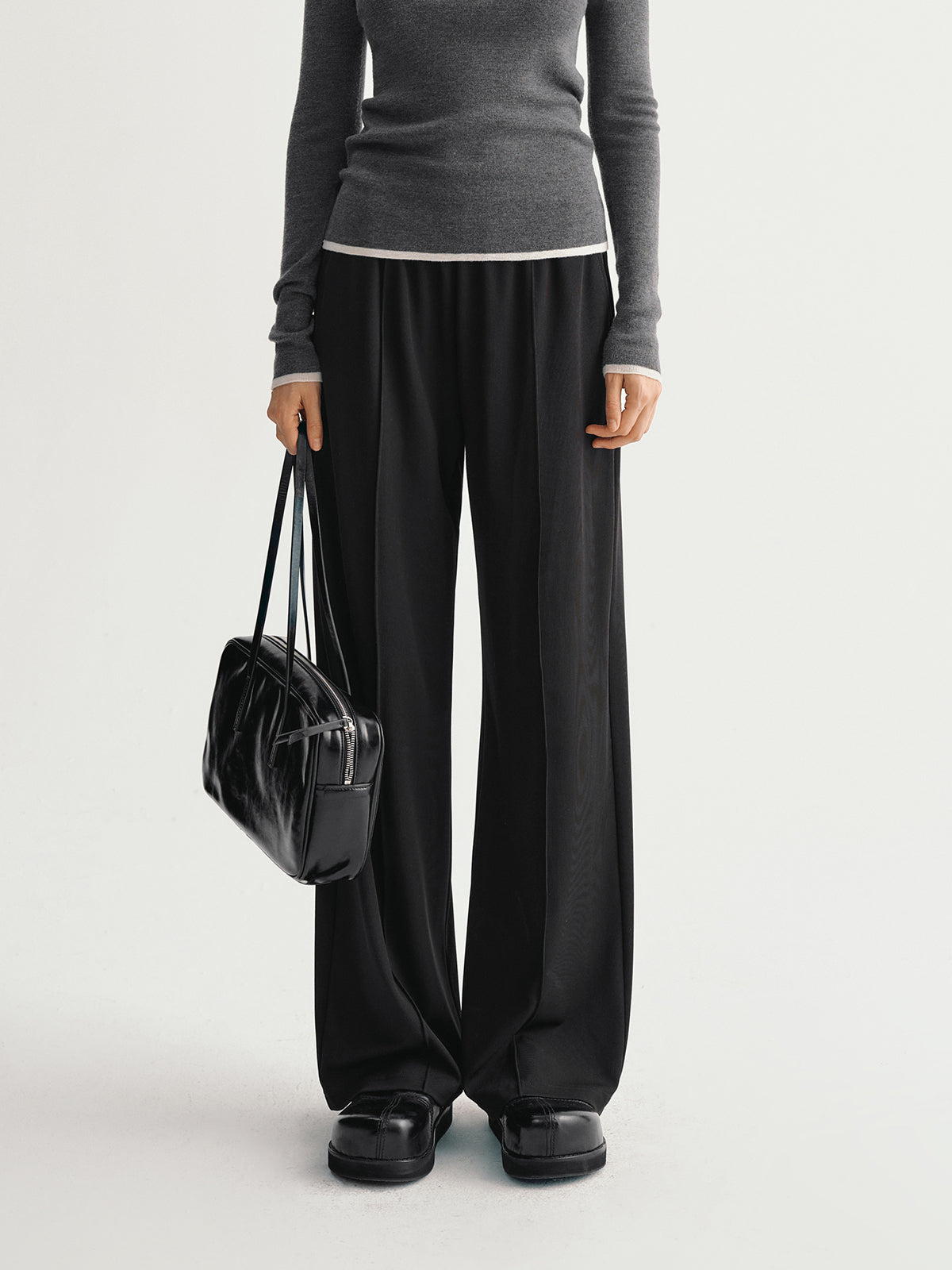 Person wearing a gray top and black wide-leg pants holding a black handbag on a light gray background