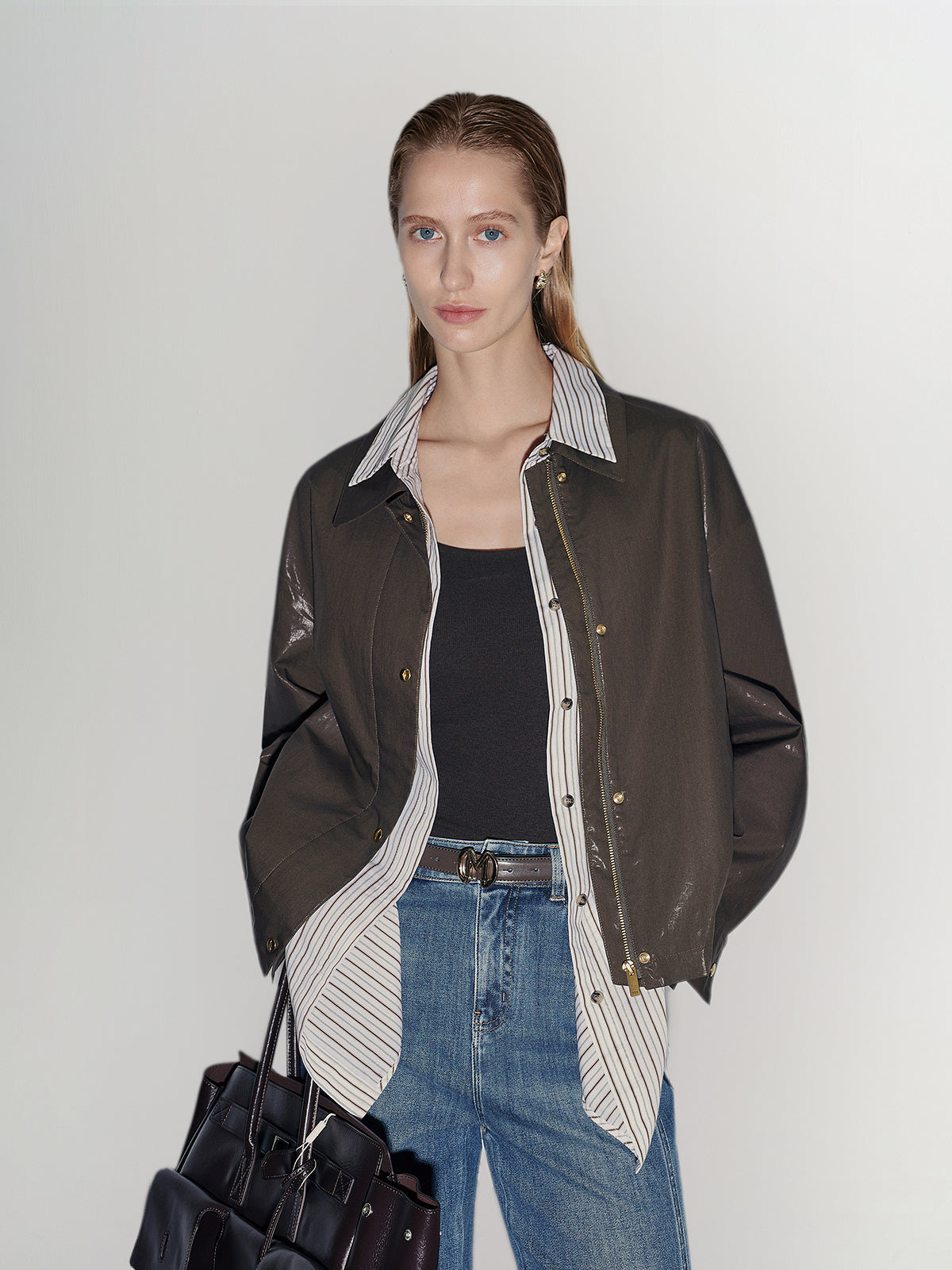 Woman wearing a brown leather jacket with a white shirt and blue jeans, holding a black handbag on a plain background.