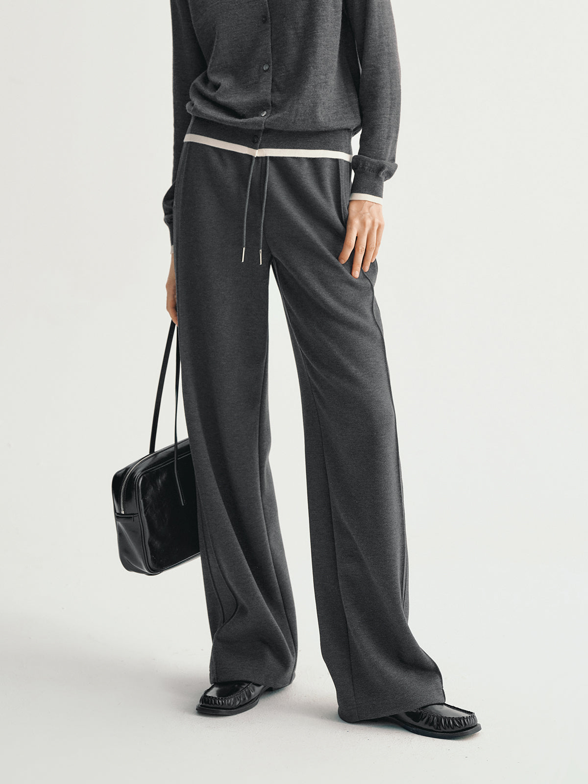 Person wearing a gray outfit with wide-leg pants and holding a black handbag on a light gray background