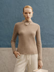 Woman wearing a beige ribbed sweater and white pants against a blue wall.