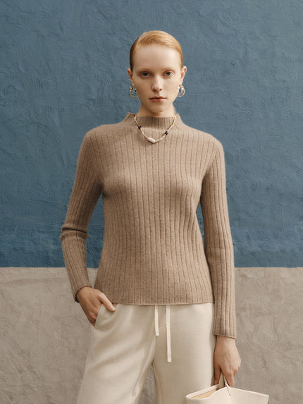 Woman wearing a beige ribbed sweater and white pants against a blue wall.