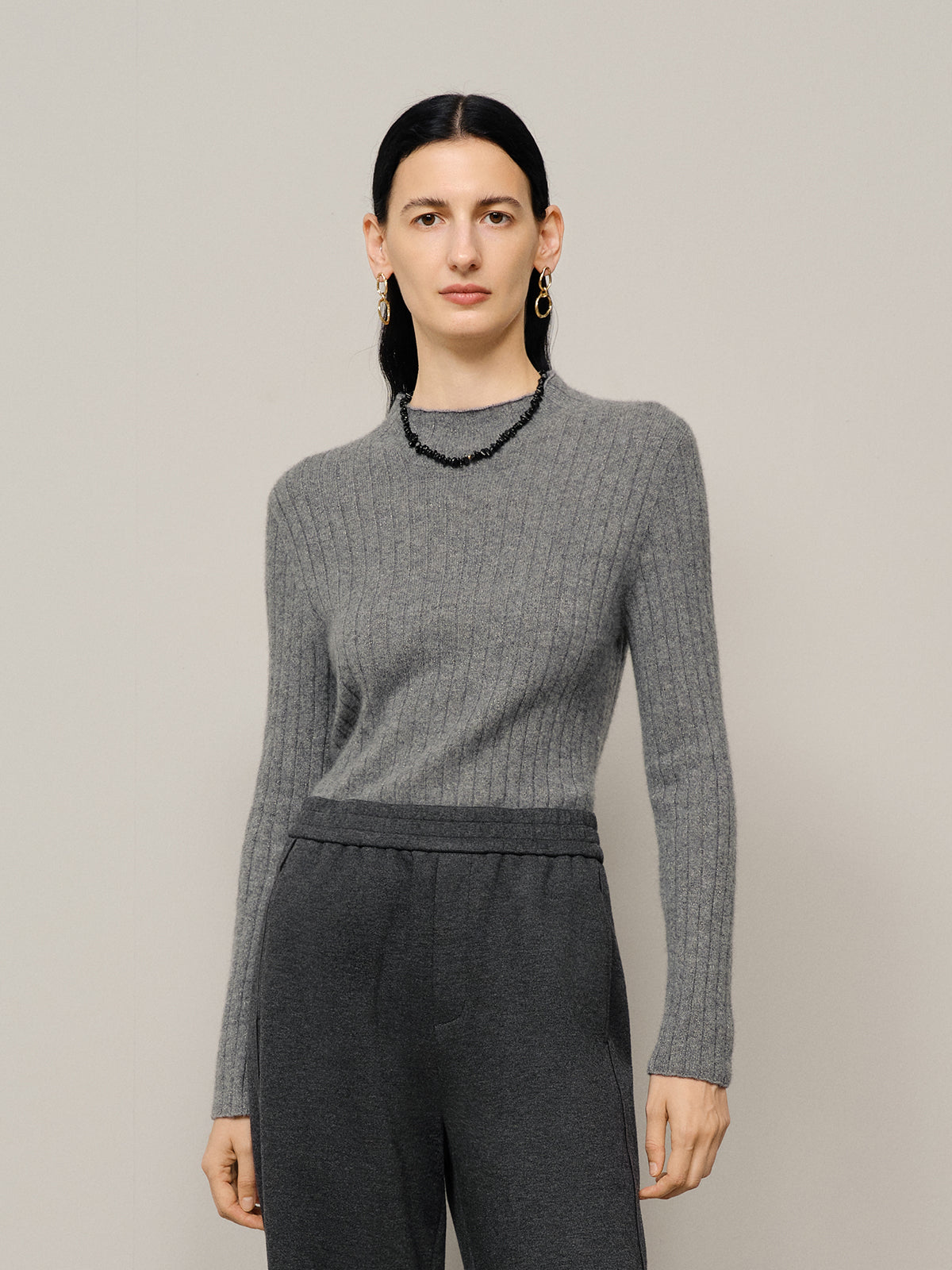 Woman wearing a gray ribbed sweater and dark pants against a plain background