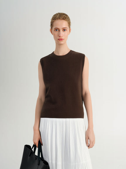 Woman wearing a brown sleeveless top and white pleated skirt against a plain background