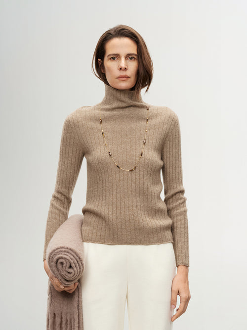 Load image 1 in gallery view. Woman wearing a beige turtleneck sweater holding a folded blanket against a plain background