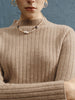 Person wearing a beige ribbed sweater and a beaded necklace against a blue background
