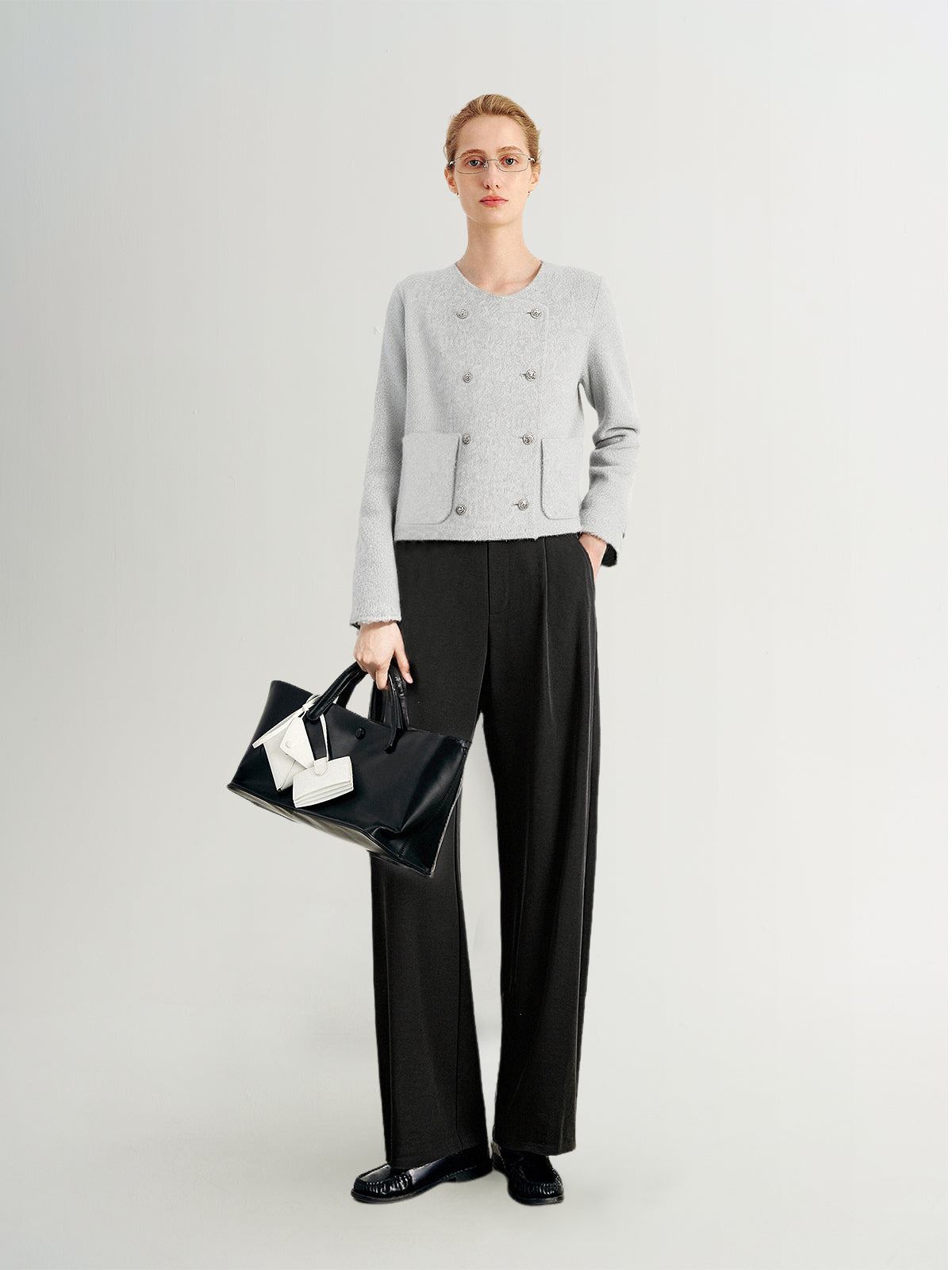 Person wearing a light gray sweater and black pants, holding a black handbag with a white bow on a white background