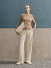 Woman wearing a beige turtleneck sweater and white pants, holding a beige handbag against a blue and gray background.