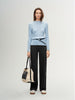 Woman wearing a light blue sweater and black pants, holding a beige and black bag on a white background