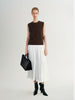 Woman wearing a brown sleeveless top, white pleated skirt, and black boots holding a black handbag on a light gray background.