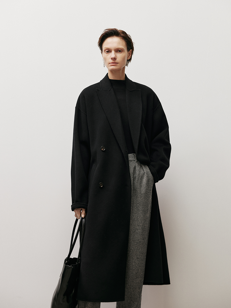 Women wearing a long black coat holding a black bag against a white background