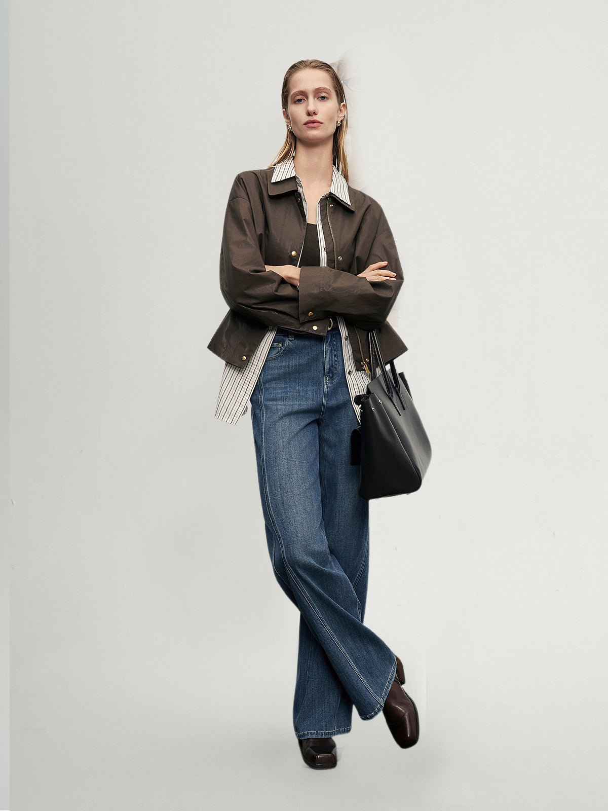 Woman wearing a brown jacket, blue jeans, and carrying a black bag on a plain background