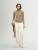 Woman wearing a beige turtleneck sweater and white pants, holding a beige scarf.
