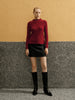 Woman wearing a red sweater, black skirt, and black boots against a textured wall.