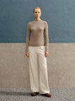 Person wearing a beige sweater and white pants against a blue and gray background