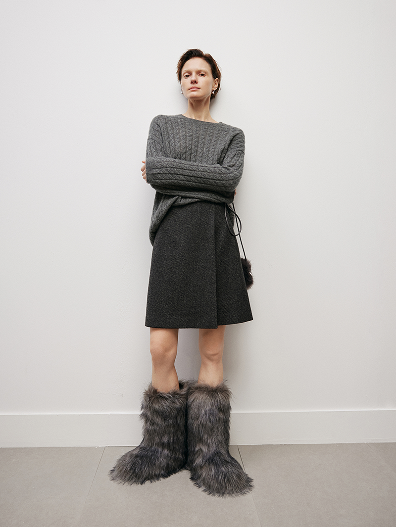 Women wearing a gray knit sweater and skirt with fur-trimmed boots against a white wall.