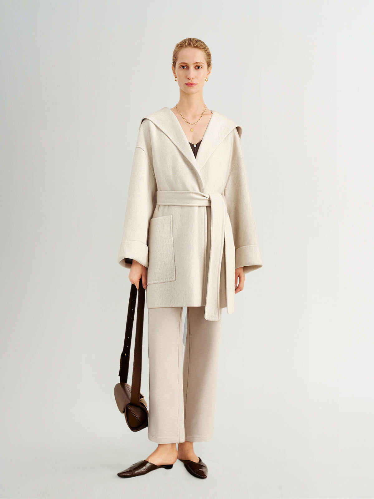 Woman wearing a beige coat and pants with a brown bag on a light gray background