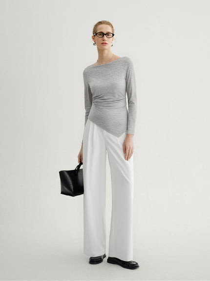 Woman wearing a gray long-sleeve top and white wide-leg pants, holding a black handbag on a light gray background.