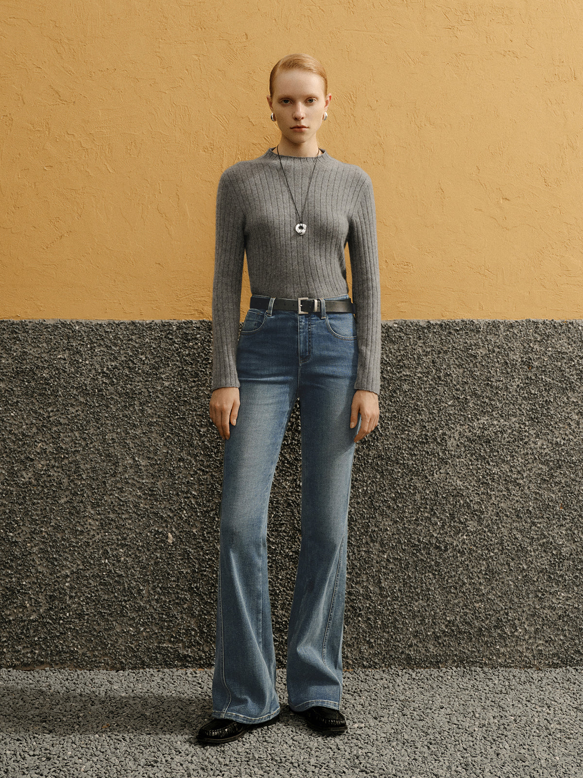Woman wearing a gray sweater and blue jeans standing against a yellow and gray wall.