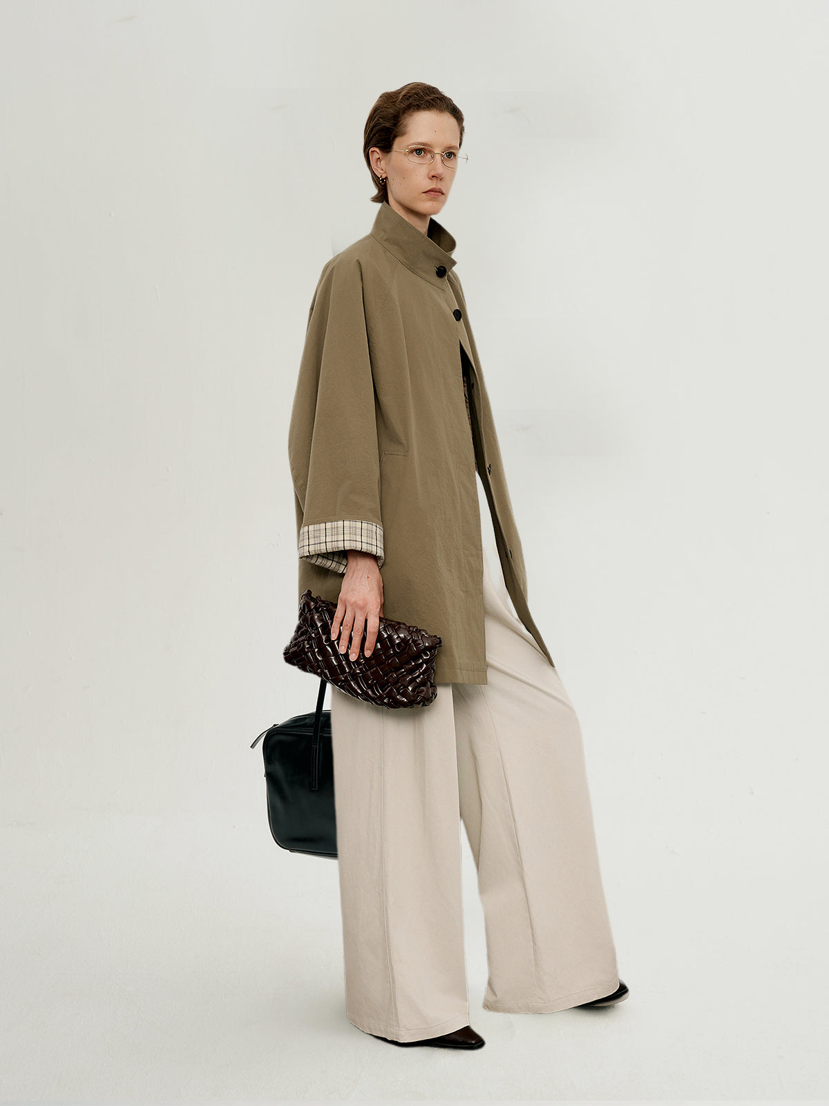Model wearing a beige coat and holding a black handbag on a white background