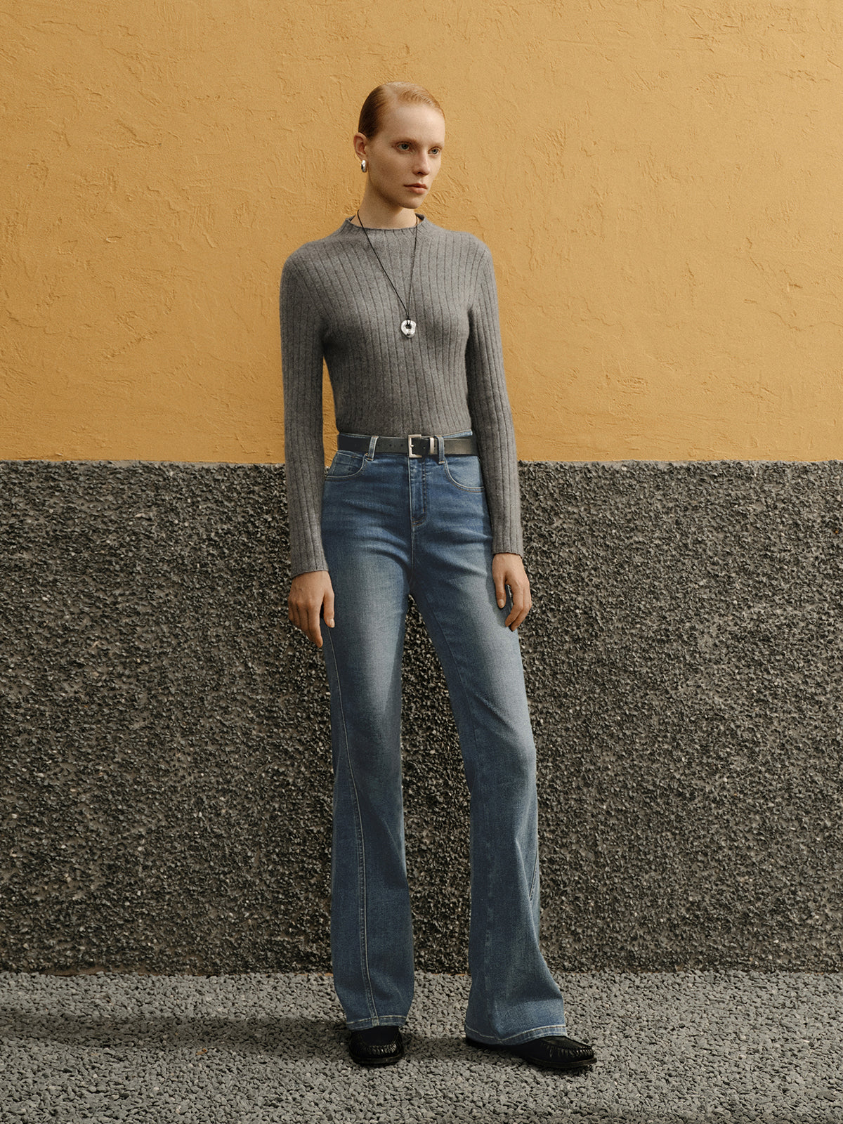 Person wearing a gray sweater and blue jeans standing against a textured wall.