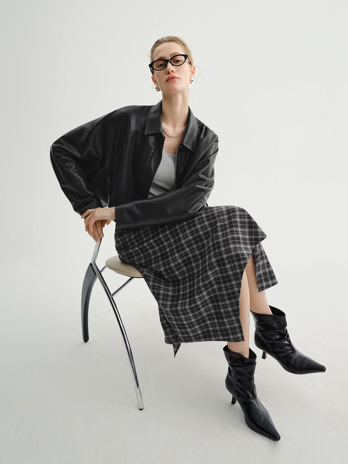 Woman sitting on a chair wearing a black leather jacket, plaid skirt, and black boots.