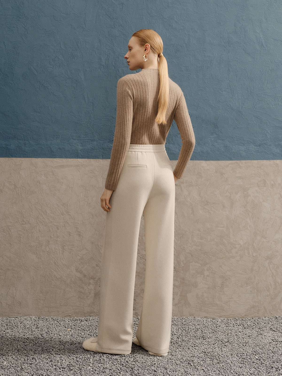 Woman in a beige sweater and white pants standing against a blue wall.