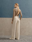 Woman in a beige sweater and white pants standing against a blue wall.