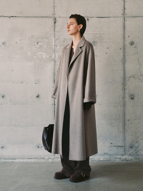 Load image 1 in gallery view. Person wearing a long gray coat against a concrete wall.
