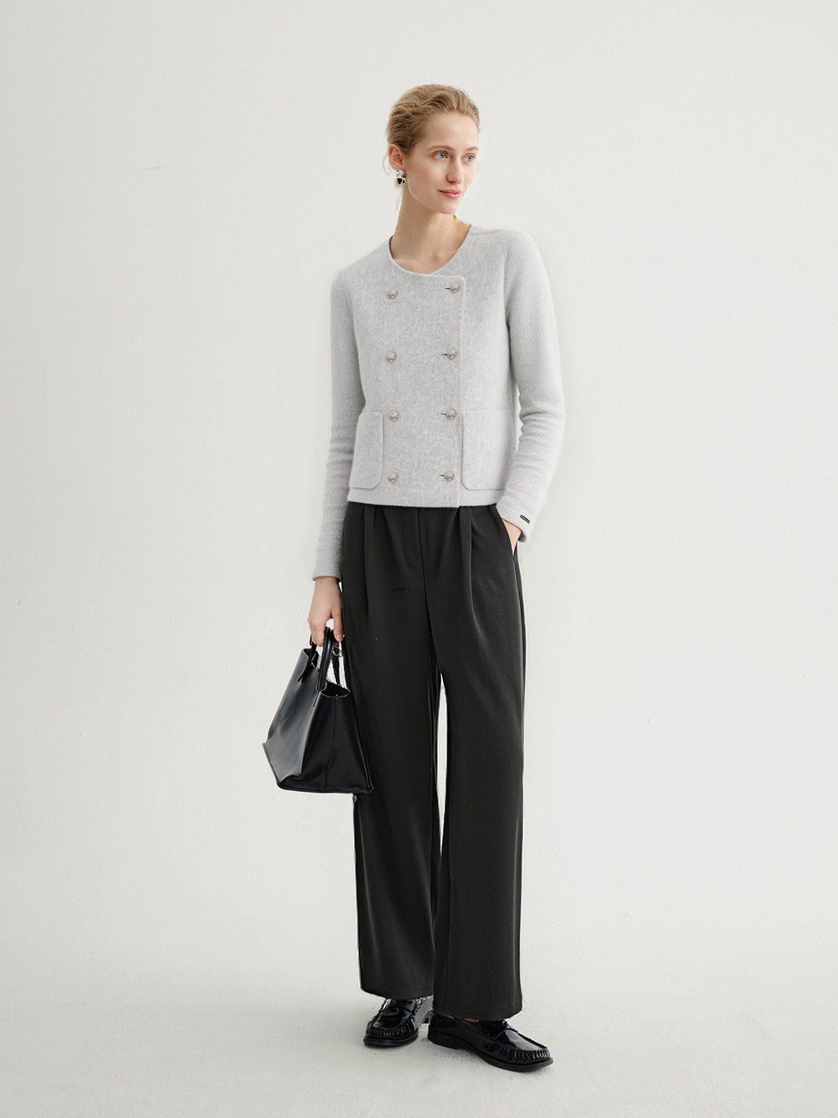 Woman wearing a gray cardigan, black pants, and holding a black handbag on a white background