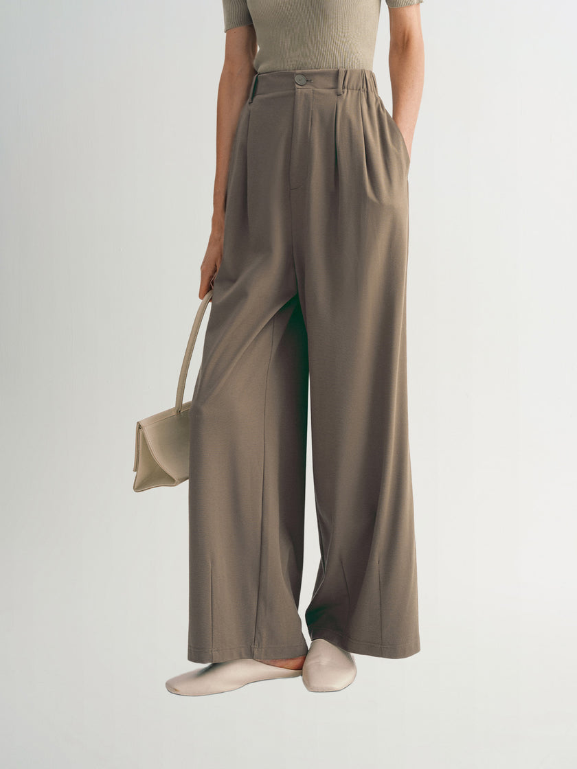A pair of wide-leg trousers in a solid color with a pleated waist and hip design, worn with a pair of white shoes and a beige handbag.