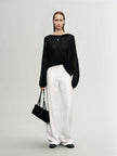 Model wearing a black Cashmere-Silk Relaxed Pullover Sweater with white trousers, holding a black handbag in a minimalist setting.