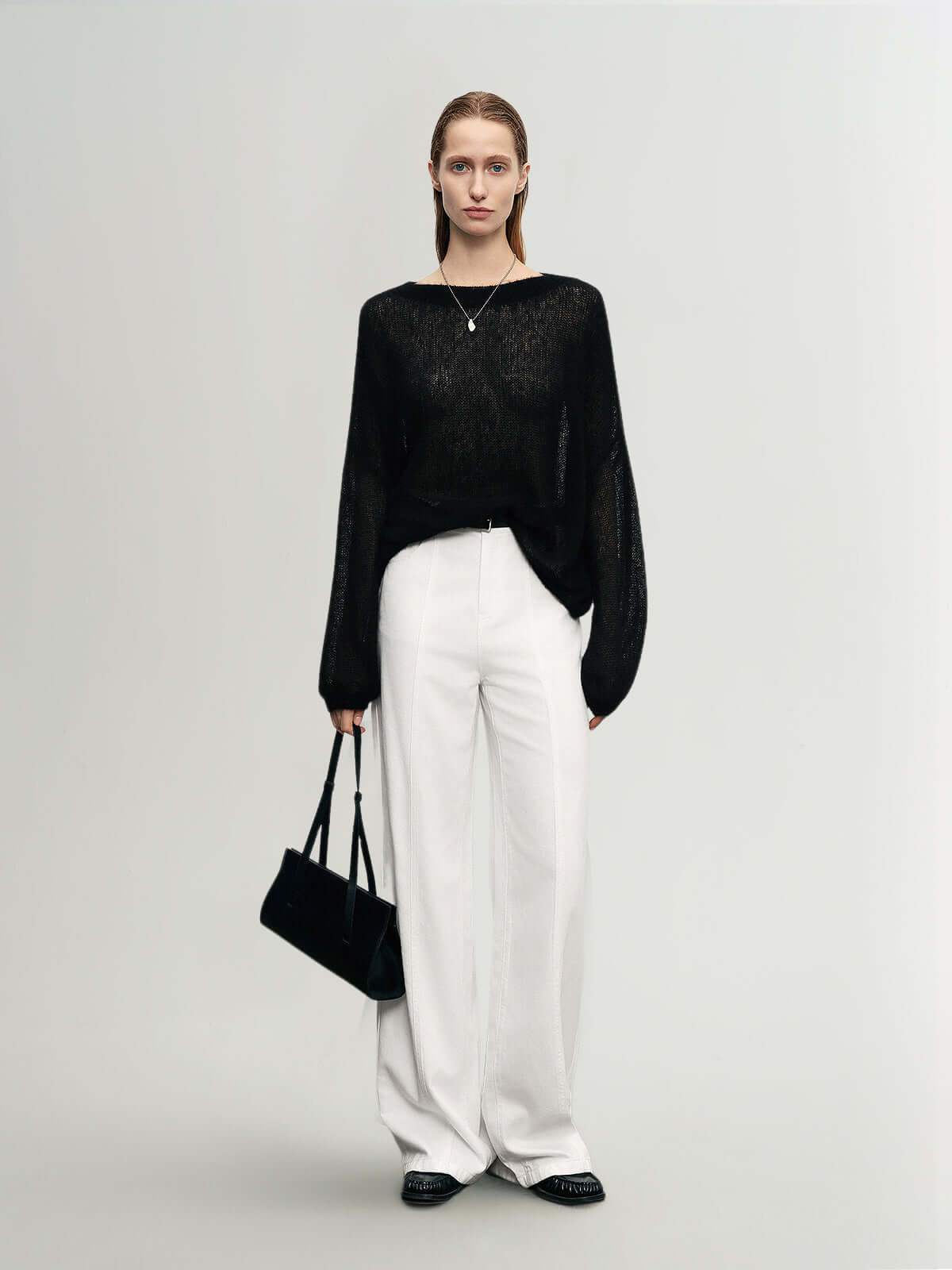 Model wearing a black Cashmere-Silk Relaxed Pullover Sweater with white trousers, holding a black handbag in a minimalist setting.