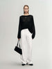 Model wearing a black Cashmere-Silk Relaxed Pullover Sweater with white trousers, holding a black handbag in a minimalist setting.